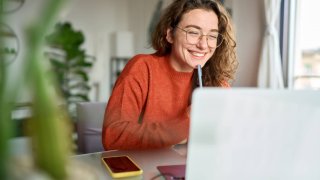 Happy young woman using laptop sitting at desk writing notes while watching webinar, studying online, looking at pc screen learning web classes or having virtual call meeting remote working from home.