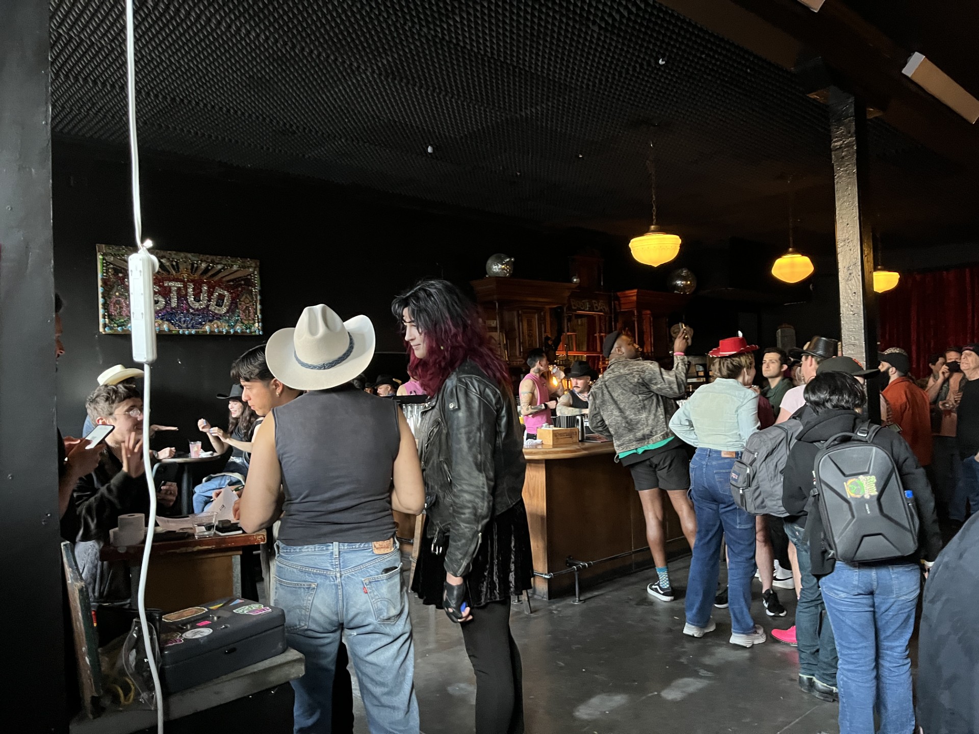 People line up in a dark bar to talk and get drinks at the bar. A sign on the wall reads 'The Stud.'