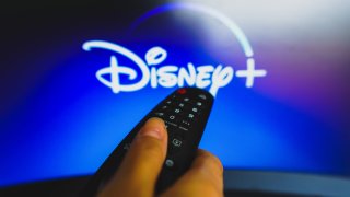 In this photo illustration, a hand holding a TV remote control in front of the Disney Plus logo on a TV screen.