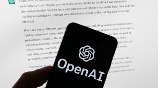 The OpenAI logo is seen on a mobile phone in front of a computer screen displaying output from ChatGPT