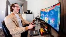 A man in a tan jacket with glasses sits at a desk, playing a game on a computer.