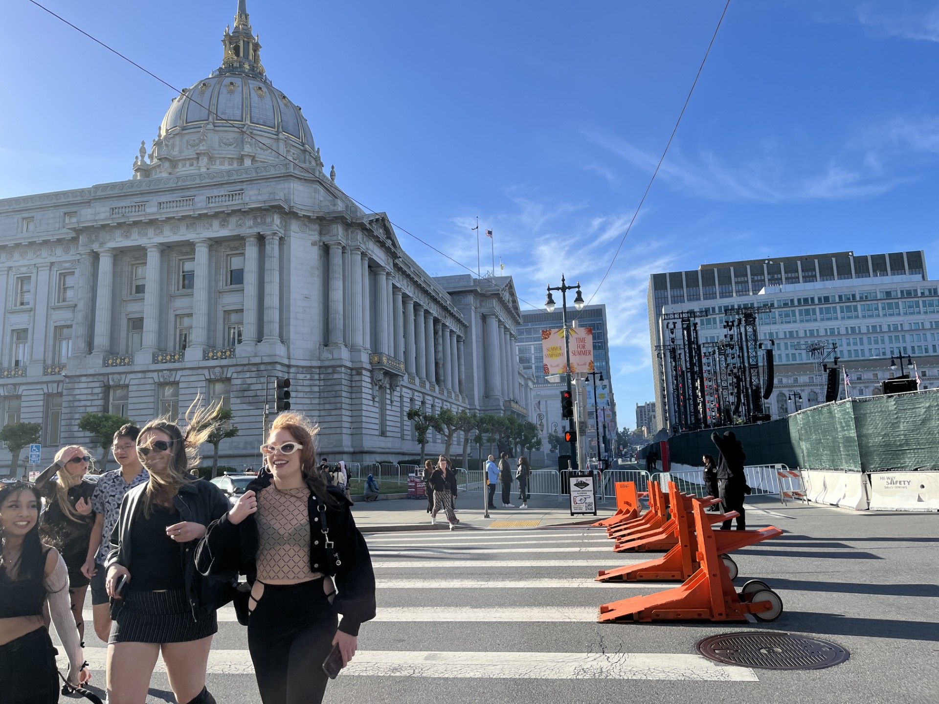 People walk across a blocked off street in San Francisco right in front of City Hall where a large stage is set up in the background.