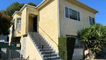 The front of a yellow duplex is pictured with a gray staircase leading up to the door on a clear, blue day.