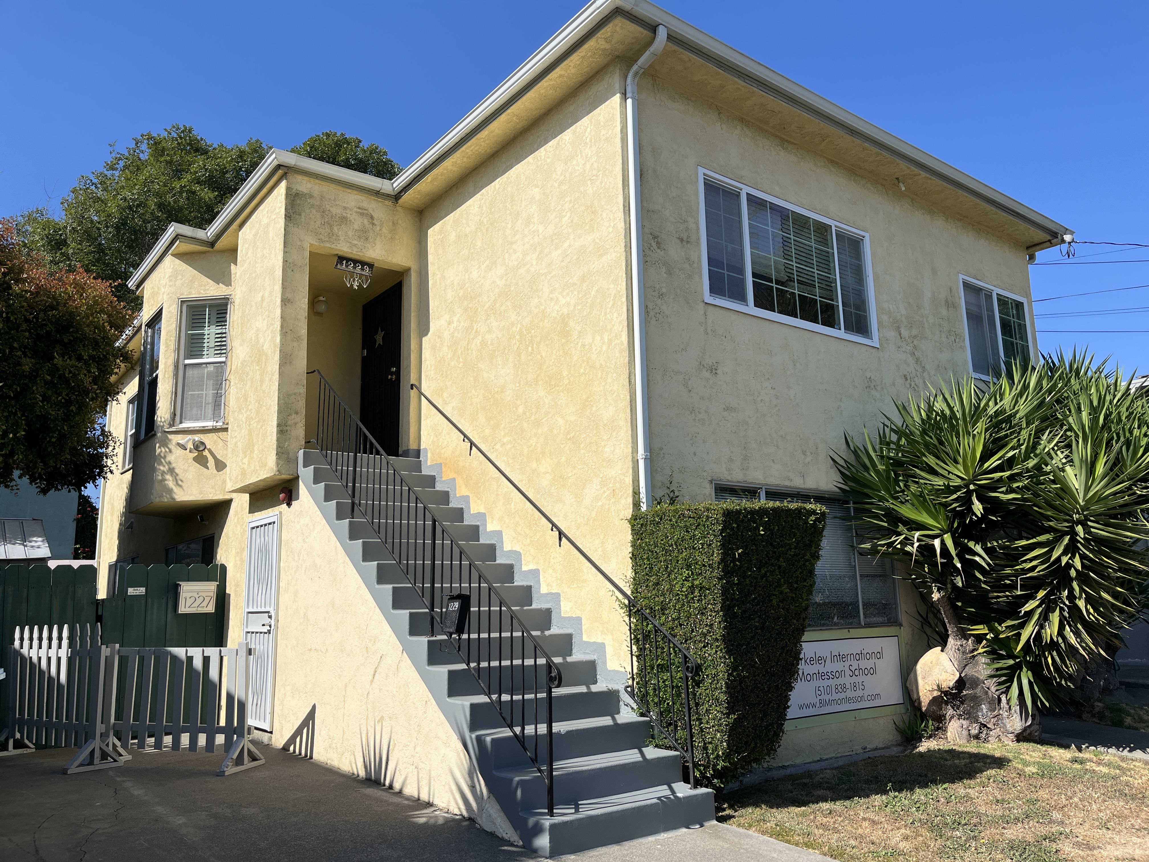 The front of a yellow duplex is pictured with a gray staircase leading up to the door on a clear, blue day.