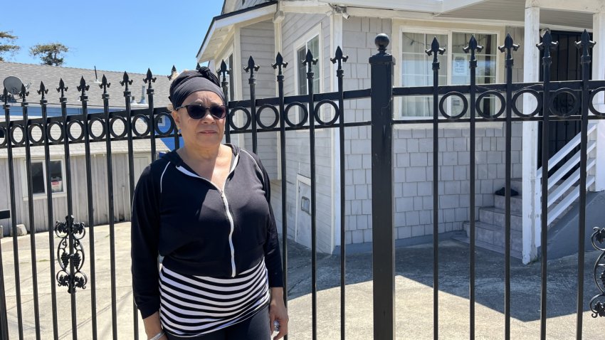 A woman in a black sweatshirt and striped shirt stands in front of a gate that protects a house behind her with white trim.