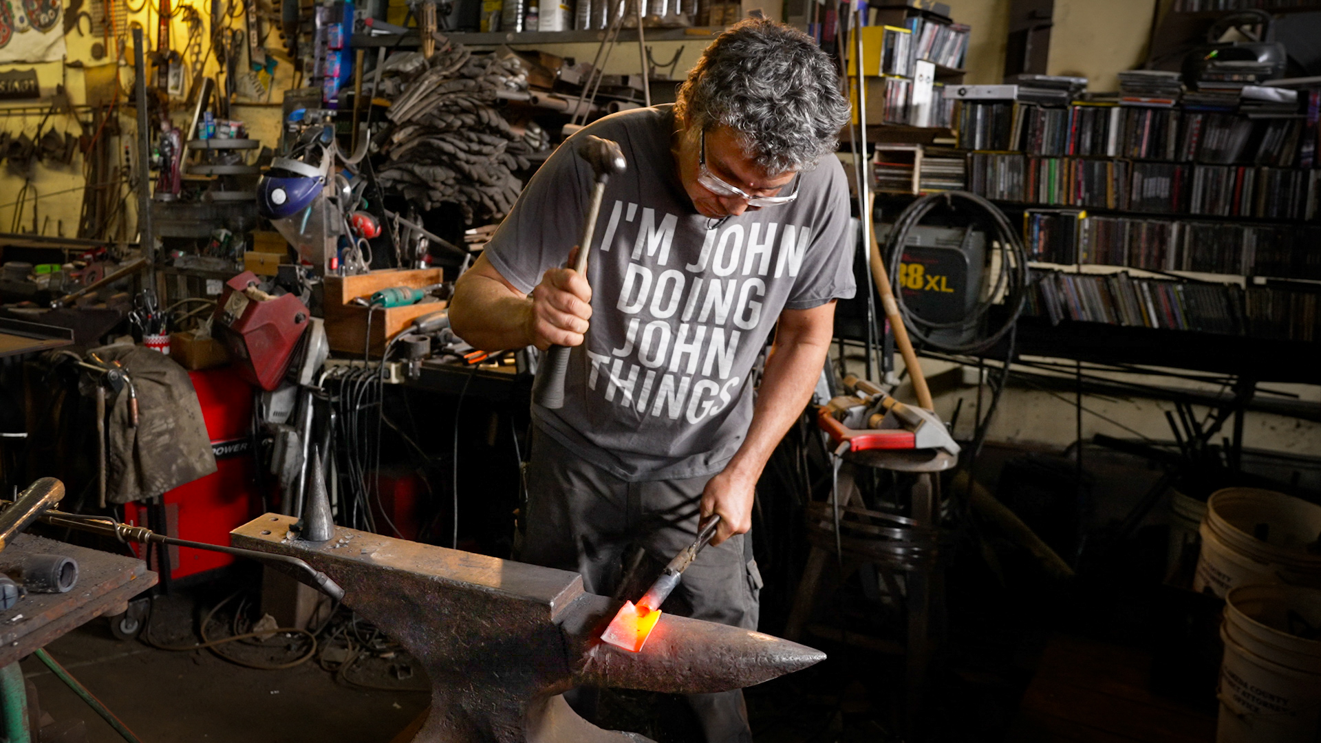 A man in a T-shirt that says "I'm John doing John things" raises a hammer to strike a glowing piece of metal on top of a large anvil.