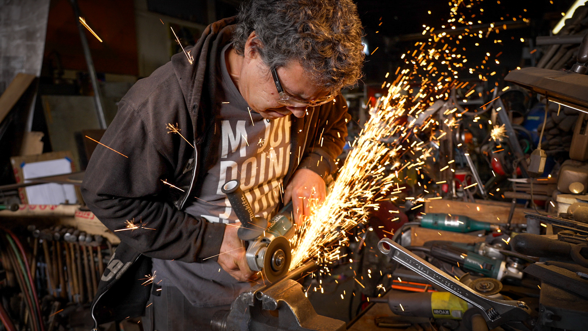 Man in glasses bent over a workbench with gold sparks flying into the air