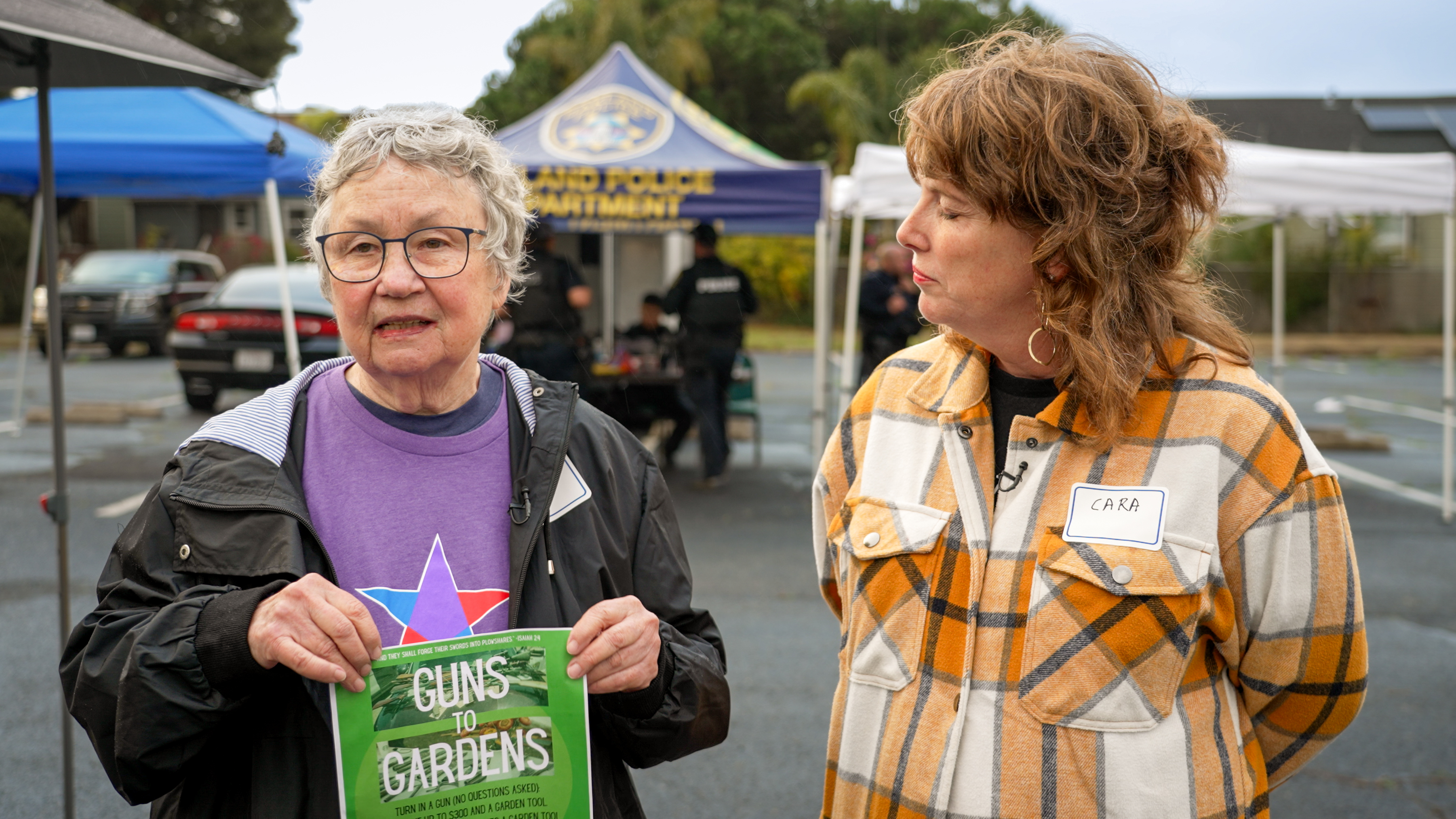 Two women speak in an interview. One holds a green poster that says "Guns to Gardens."