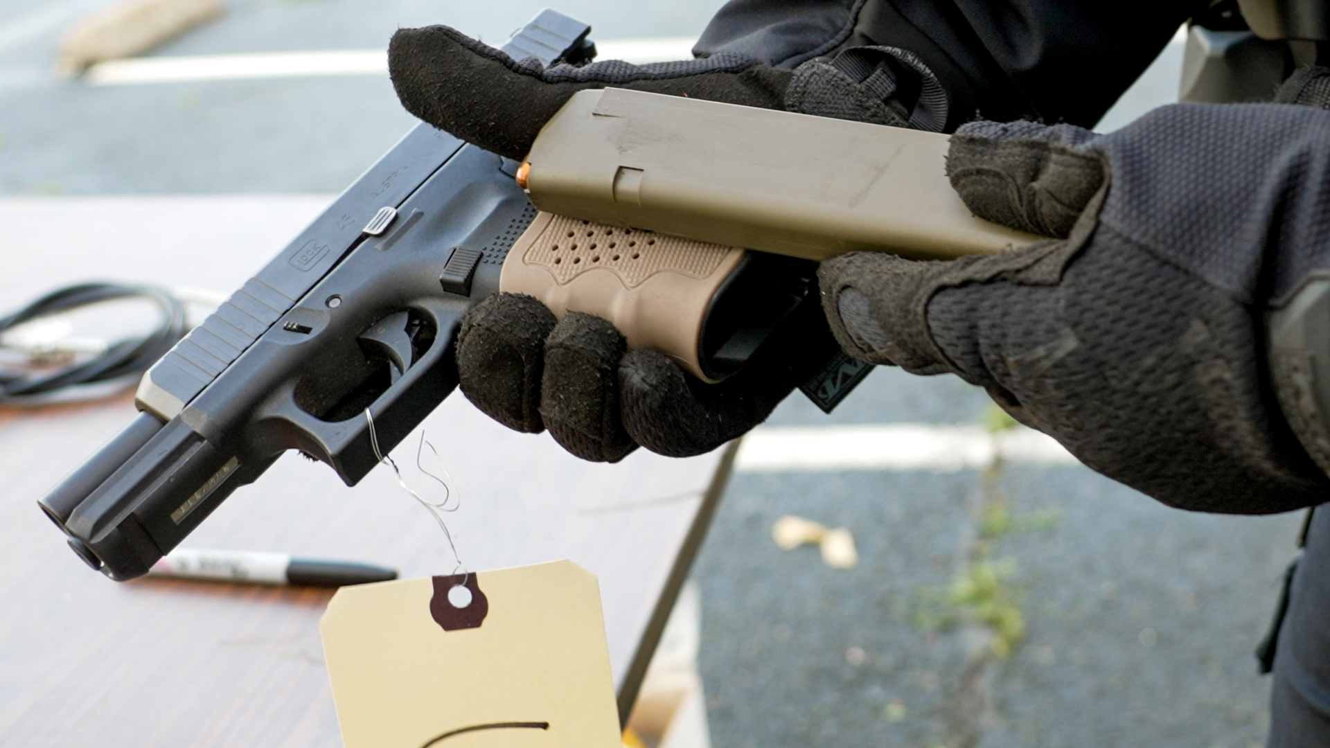 A handgun with an olive green rubber grip is held in gloved hands along with an olive green plastic magazine loaded with ammunition.