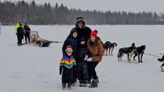 Patrice Poltzer poses with her husband and three boys for a family photo in Finland with a sled and dogs in the snowy backdrop.