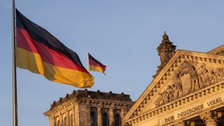 Two German flags fly in front of and on top of the Reichstag building at sunset.