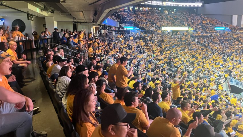 Fans inside Chase Center wearing yellow Golden State Warriors shirts cheer.