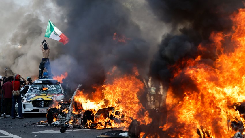 LOS ANGELES, LOS ANGELES – JUNE 08: A protestor holds up a Mexican flag as burning cars line the street on June 08, 2025 in Los Angeles, California. Tensions in the city remain high after the Trump administration called in the National Guard against the wishes of city leaders following two days of clashes with police during a series of immigration raids.  (Photo by Mario Tama/Getty Images)